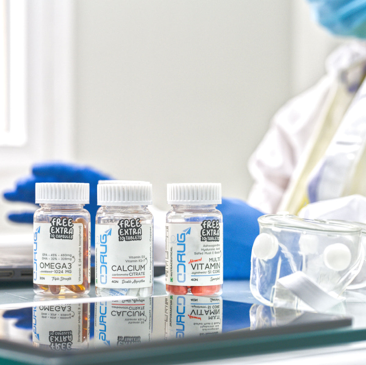 Three small supplement bottles labeled DRUG Rocket Vitamins are arranged on a reflective glass surface in a bright clinical or laboratory setting, with a person in a white coat and blue gloves blurred in the background.