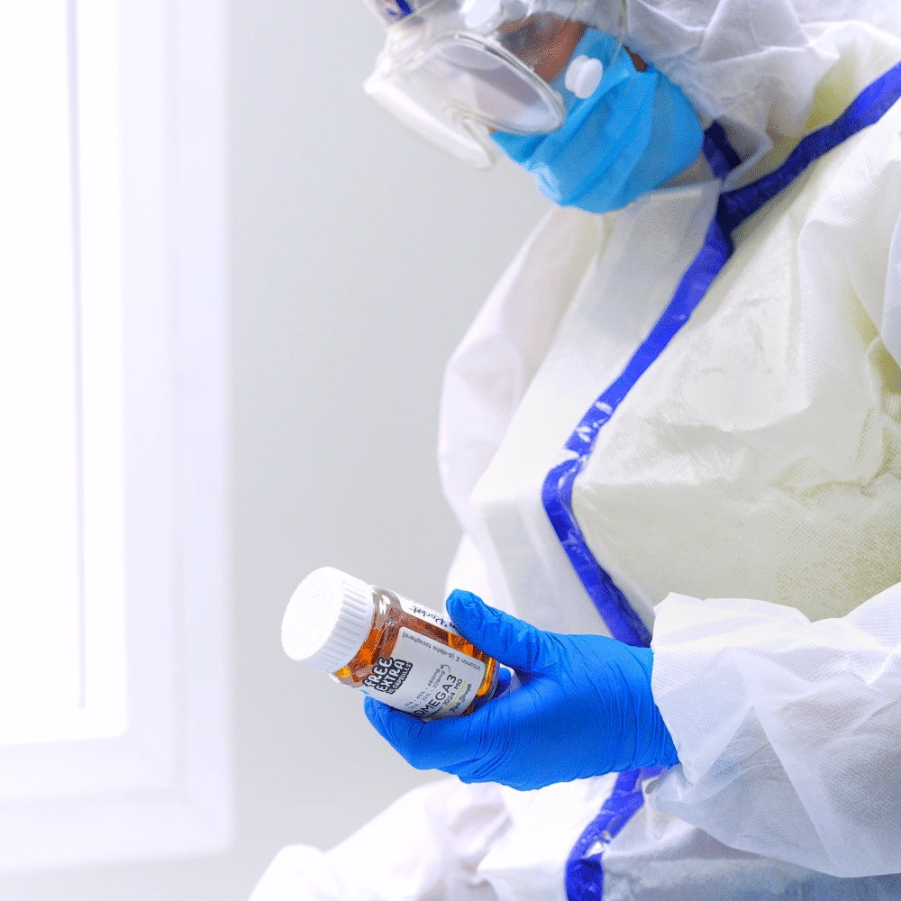 A person in a white protective suit with blue trim, blue gloves, and a face shield holds an Drug Rocket Omega-3 bottle with a white cap in a bright clinical setting.