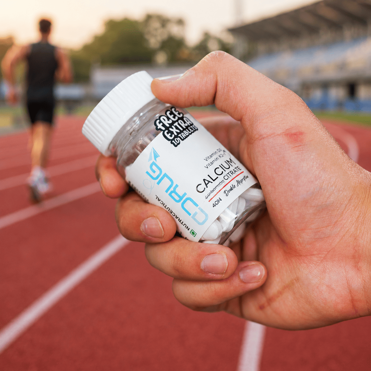 Hand holding a supplement bottle of Drug Rocket 'Calcium Citrate' on a running track. This is a Drug Rocket Calcium Citrate with Vitamin D3 & Vitamin K2-7, including Zinc, Magnesium, Copper. With 10 extra free tablets.